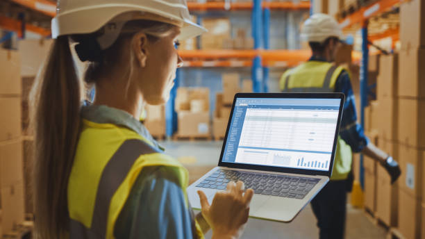 istock Professional Female Worker Wearing Hard Hat Holds Laptop Computer with Screen Showing Inventory Checking Software in the Retail Warehouse full of Shelves with Goods. Over the Shoulder Side View 1284193250