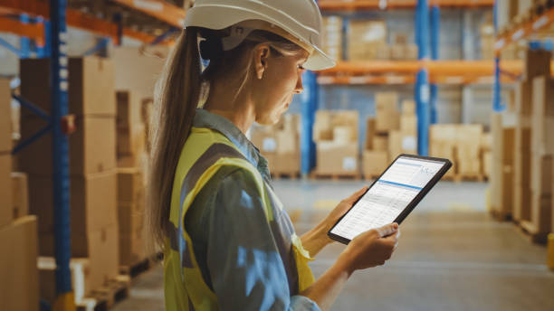istock Professional Female Worker Wearing Hard Hat Uses Digital Tablet Computer with Inventory Checking Software in the Retail Warehouse full of Shelves with Goods. Delivery, Distribution Center. 1284193348
