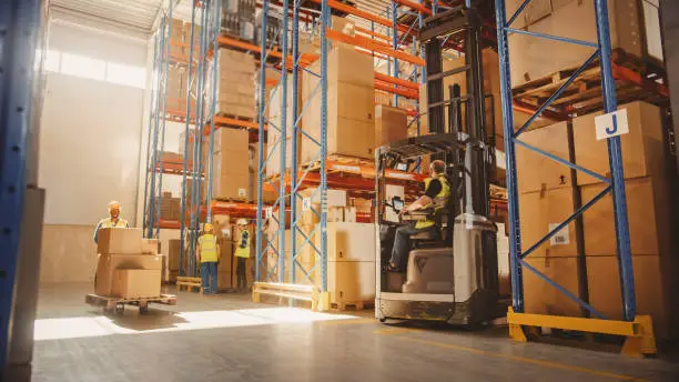Retail Delivery Warehouse full of Shelves with Goods in Cardboard Boxes, Workers Scan and Sort Packages, Move Inventory with Pallet Trucks and Forklifts. Product Distribution and Delivery Logistics.