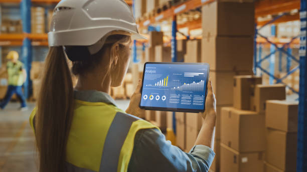 istock Professional Female Worker Wearing Hard Hat Uses Digital Tablet Computer with Screen Showing Analysis Software in the Retail Warehouse full of Shelves with Goods. Over the Shoulder View 1284193383