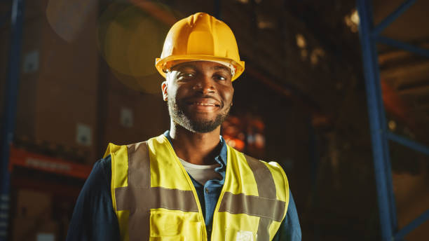 travailleur professionnel beau et heureux portant le gilet de sécurité et le chapeau dur souriant charmant sur l’appareil-photo. dans le grand entrepôt de fond avec des étagères pleines de marchandises de livraison. portrait de plan rapproché moyen - charmingly photos et images de collection