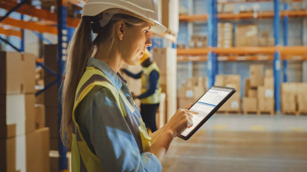istock Professional Female Worker Wearing Hard Hat Uses Digital Tablet Computer with Inventory Checking Software in the Retail Warehouse full of Shelves with Goods. Delivery, Distribution Center. 1284193443