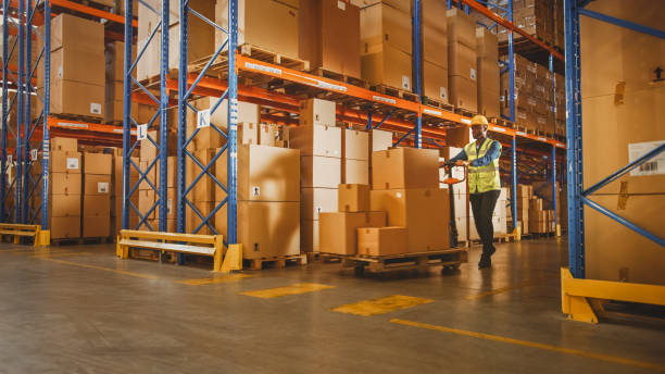 istock Worker Moves Cardboard Boxes using Hand Pallet Truck, Walking between Rows of Shelves with Goods in Cardboard Boxes in Retail Warehouse. Product Delivery Distribution Logistics Center. 1284193447