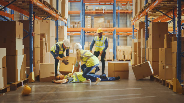 istock Warehouse Worker Has Work Related Accident Falls while Trying to Pick Up Cardboard Box from the Shelf. Colleagues Call for Help and Medical Assistance. Injury at Work. 1284193466