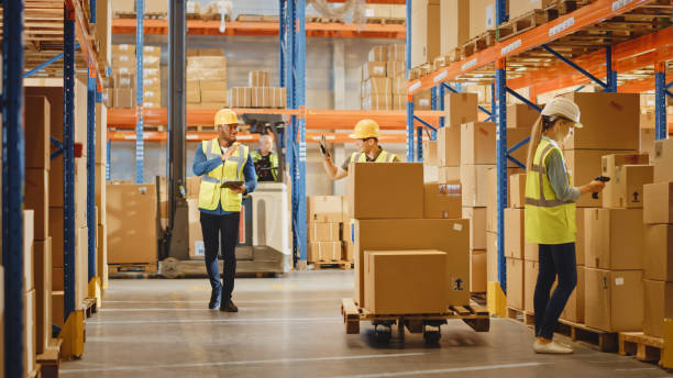 istock Handsome Male Worker Wearing Hard Hat Holding Digital Tablet Computer Walking Through Retail Warehouse full of Shelves with Goods, Greeting Workers. Working in Logistics and Distribution Center. 1284193509