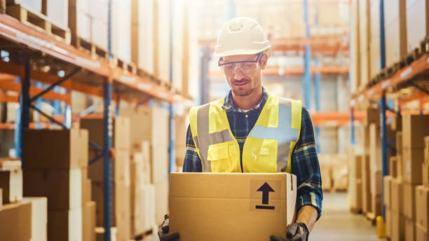 istock Handsome Male Worker Wearing Hard Hat Holding Cardboard Box Walking Through Retail Warehouse full of Shelves with Goods. Working in Logistics and Distribution Center. Front Shot 1284193521