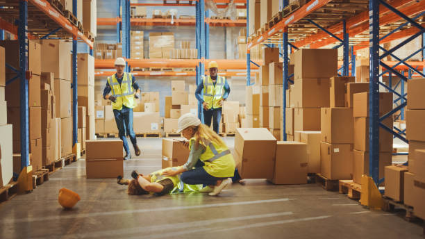 istock Warehouse Worker Has Work Related Accident Falls while Trying to Pick Up Cardboard Box from the Shelf. Colleagues Call for Help and Medical Assistance. Injury at Work. 1284193548