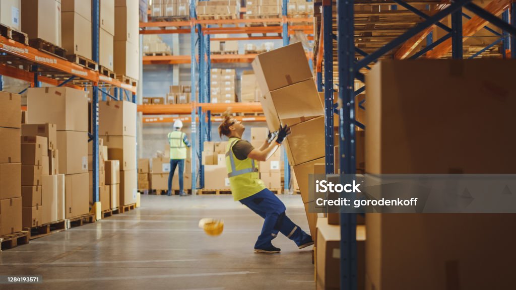 Shot of a Warehouse Worker Has Work Related Accident. He is Falling Down BeforeTrying to Pick Up Heavy Cardboard Box from the Shelf. Hard Injury at Work. Occupation Stock Photo