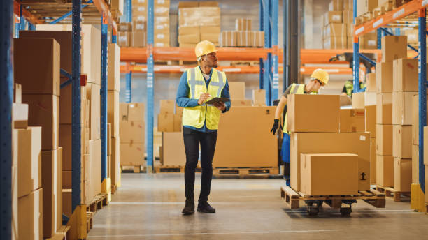 istock Handsome Male Worker Wearing Hard Hat Holding Digital Tablet Computer Walking Through Retail Warehouse full of Shelves with Goods. Professional People Working in Logistics and Distribution Cente 1284193576
