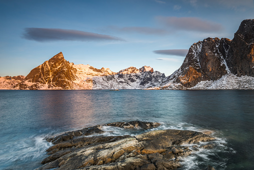 Ice sheets on dark rock with lake and snow-clad steep mountains in the Reinefjord on the Lofoten islands in Norway in golden morning light on clear winter day