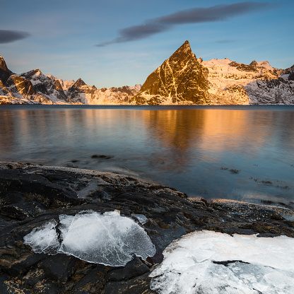 Ice sheets on dark rock with lake and snow-clad steep mountains in the Reinefjord on the Lofoten islands in Norway in golden morning light on clear winter day