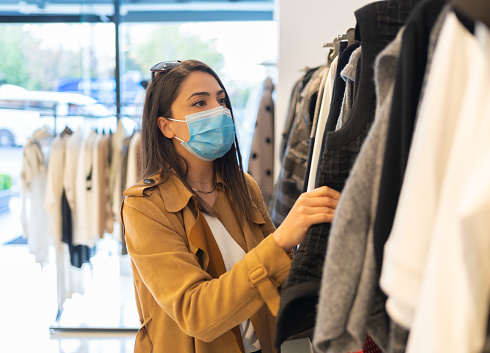 A beautiful young stylish woman with protective face mask is choosing trendy dress in the clothing store during pandemic