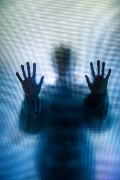 Silhouette of depressed abused woman with hands pressed against glass Colour backlit image of the silhouette of a woman with her hands pressed against a glass window. The silhouette is distorted, and the arms elongated, giving an alien-like quality. The image is sinister and foreboding, with an element of horror. It is as if the 'woman' is trying to escape from behind the glass. Horizontal image with copy space. people trafficking stock pictures, royalty-free photos & images