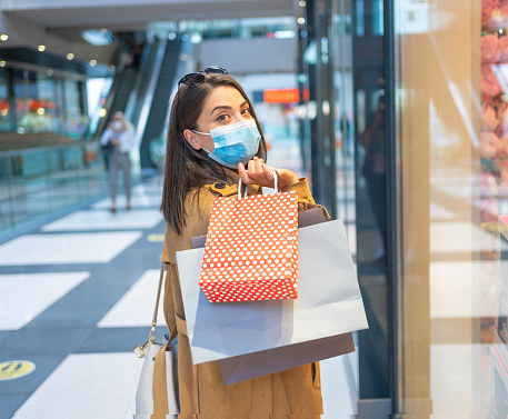 Happy and beautiful young woman with protective face mask is holding shopping paper bags while  enjoying at the shopping mall