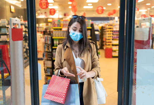 istock Young beautiful woman with protective face mask is cleaning her hands with alcohol disinfectant spray after buying cosmetics from beauty shop at the shopping mall during coronavirus 1284202478