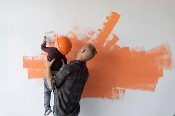 Photo of repairs his house, the father keeps his son and he helps him paint the wall with a roller,