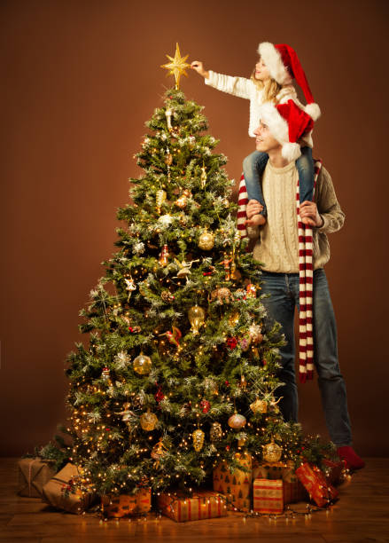 クリスマスファミリーはクリスマスツリーを飾ります。サンタハットの子供は上に星を置きます。屋内肖像画と一緒に娘と幸せな父。クリスマスツリーのライトとボーブルの暗闇 - christmas stack gift carrying ストックフォトと画像