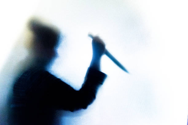 Violent silhouette of person wielding a knife behind frosted glass window Backlit image of the silhouette of a person, probably a woman, wielding a sharp knife in an aggressive way. The silhouette is distorted, and the arms elongated, giving an alien-like quality. The image is sinister and foreboding, with an element of horror. The image conveys a domestic violence, knife crime theme. Horizontal image with copy space. serial killings photos stock pictures, royalty-free photos & images