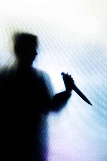 Violent silhouette of person wielding a knife behind frosted glass window Backlit image of the silhouette of a person, probably a woman, wielding a sharp knife in an aggressive way. The silhouette is distorted, and the arms elongated, giving an alien-like quality. The image is sinister and foreboding, with an element of horror. The image conveys a domestic violence, knife crime theme. Horizontal image with copy space. knife crime photos stock pictures, royalty-free photos & images