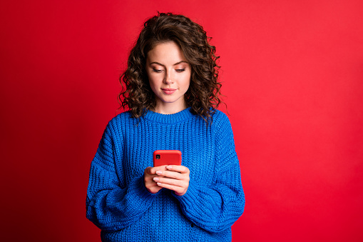 Photo of cute enigmatic charming lady wavy hairstyle hold telephone, calm confident look screen smile relaxing wear blue knitted sweater pullover isolated red color background