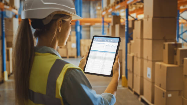 istock Professional Female Worker Wearing Hard Hat Uses Digital Tablet Computer with Inventory Checking Software in the Retail Warehouse full of Shelves with Goods. Delivery, Distribution Center. Side View 1284212354