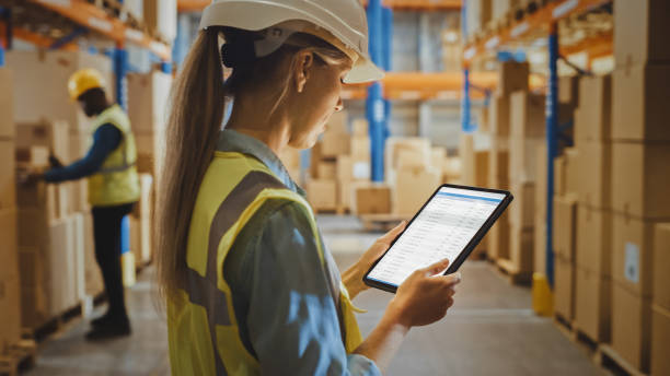 istock Professional Female Worker Wearing Hard Hat Uses Digital Tablet Computer with Inventory Checking Software in the Retail Warehouse full of Shelves with Goods. Delivery, Distribution Center. 1284212361
