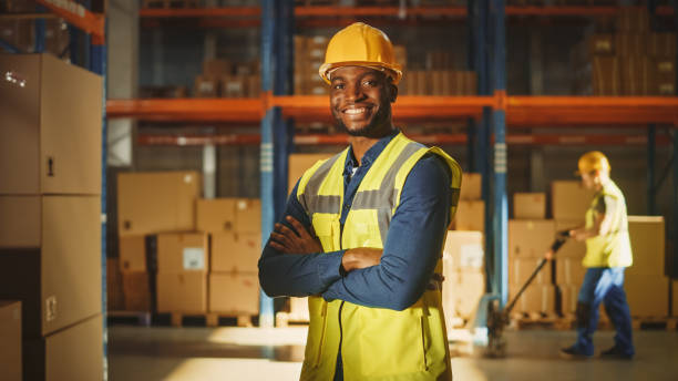 trabalhador profissional bonito e feliz vestindo colete de segurança e chapéu duro sorrindo com braços cruzados na câmera. no armazém grande fundo com prateleiras cheias de produtos de entrega. retrato médio - work wear factory people occupation - fotografias e filmes do acervo