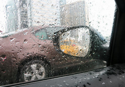 A heavy rainstorm caused a traffic jam on the road. The car's side window and mirror are drenched in raindrops. View from the car window in the rain.