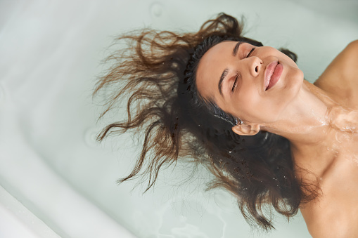 Close up of smiling lady with closed eyes floating in water while taking bath