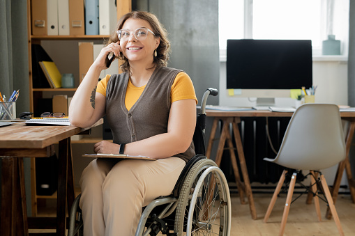 Happy young female agent or secretary in wheelchair sitting by table in modern office and consulting clients on mobile phone by workplace