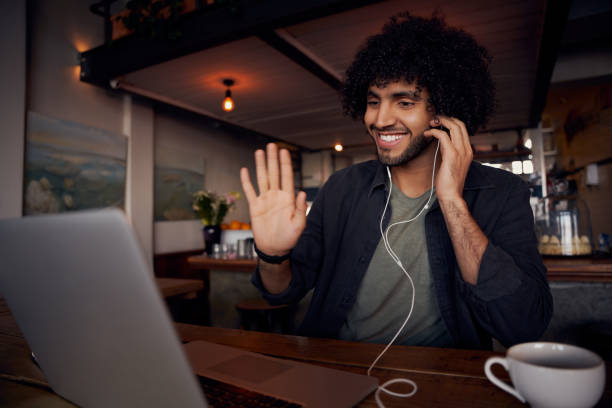 istock Handsome young man waving hand at laptop screen during video call with earphone in cafe 1284228565