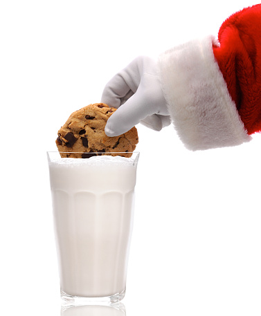 Santa Claus dunking a chocolate chip cookie into a glass of milk over a white background.