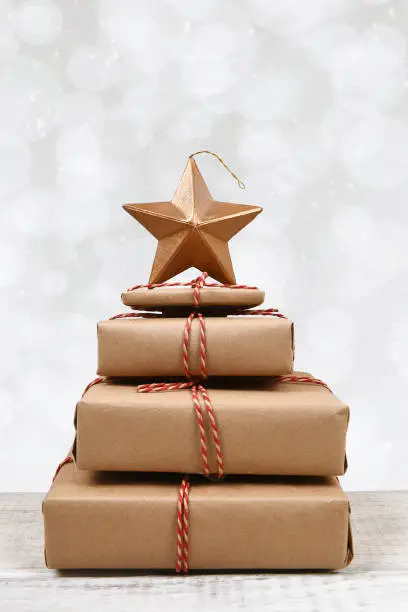 Plain brown paper wrapped gifts tied with twine and stacked in the shape of a Christmas tree with star, silver bokeh background.