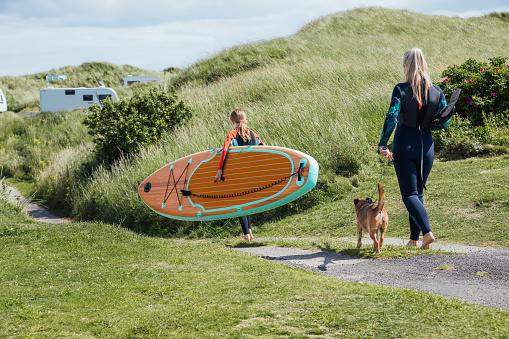 An unrecognisable mother and daughter walking down a path with their dog in a caravan park. They are wearing wetsuits while the teenage girl is carrying a paddleboard and her mother is walking the dog on a leash and is carrying the paddles. It's a beautiful summers day in the North East of England.