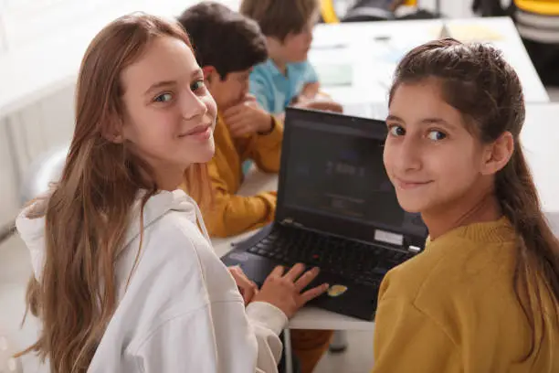 Happy teenage girls smiling to the camera while working on a school project together, using laptop