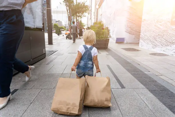 Photo of Small cute blond child is carring a lot of shopping bags at the street after black Friday sale. View from the back. Toddler is happy and feels she is a shopoholic.