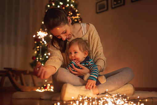 Beautiful young mother holding her son in her lap, sitting on the floor next to nicely decorated Christmas tree, playing with sparklers and christmas lights