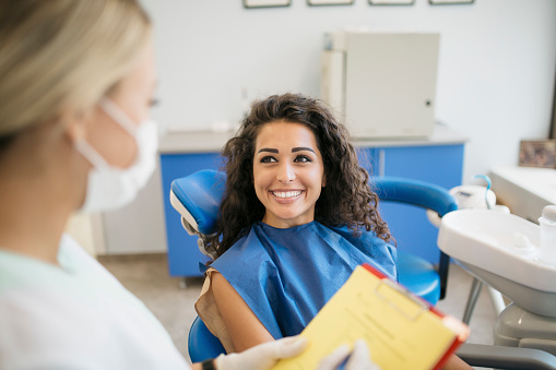 Happy young Caucasian woman smiling and sitting in a dentist's office, explaining her problems while her female dentist is listening and taking notes about her oral health