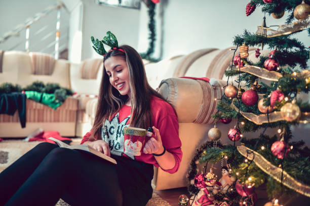 istock Cute Female With Christmas Antlers Drinking Tea And Reading Book Under Tree 1284263694