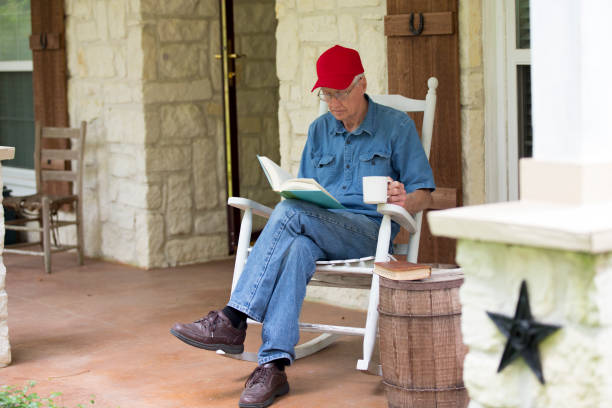 Active senior man sitting in white rocker on porch.  He wears denim and a red hat.  He is reading a book.