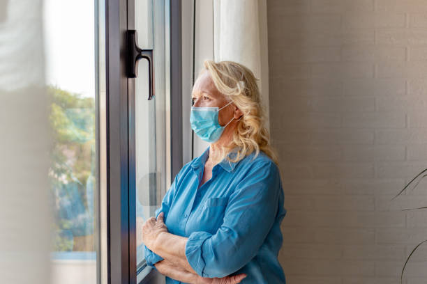 istock Senior woman at home with face mask, looking through the window 1284273100