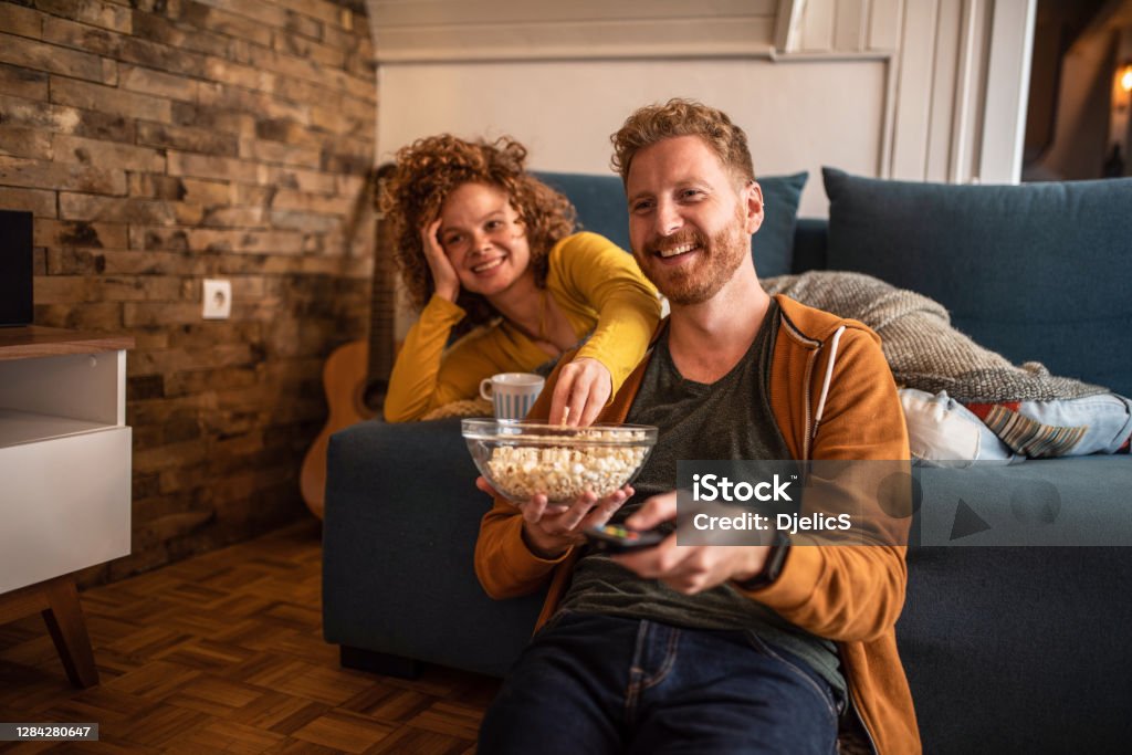 Young couple watching a movie at home. Cute young ginger couple watching tv in the living room. Movie Stock Photo