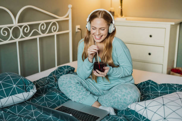 jeune femme dans des pyjamas sur un appel vidéo avec des amis - using computer bedroom one woman only cheerful photos et images de collection