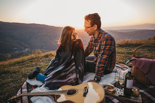 Romantic young couple with acoustic guitar sitting on picnic blanket, looking each other and smiling, and enjoying  beautiful sunset over mountains