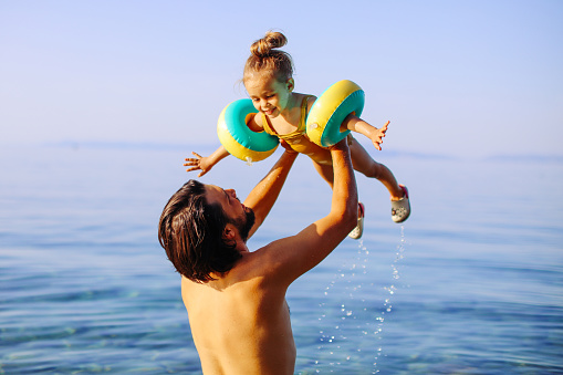 Father is throwing his daughter into the water with lots of splashing. Cute little girl having fun with parents in sea