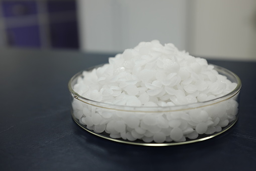 Selective focus of sodium or potassium hydroxide white chemical compound pellets or prills in a petri dish in black background.