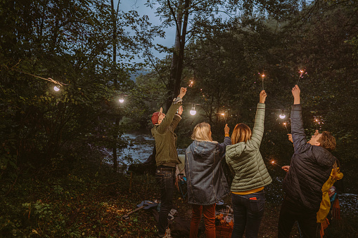 Photo of a group of friends celebrating New Year's by throwing an outdoor party, on a cold evening on a riverbank; lightning sparklers, dancing, and enjoying each other's company.