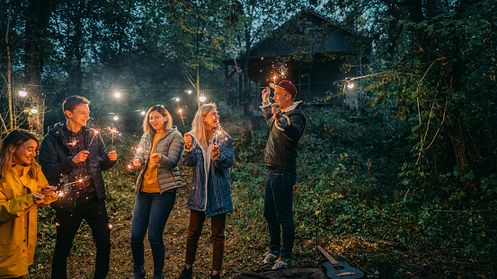 Photo of a group of friends celebrating New Year's by throwing an outdoor party, on a cold evening in the forest; lightning sparklers, dancing, and enjoying each other's company.