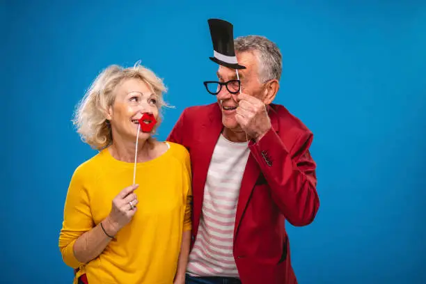 Front view close-up of playful Caucasian woman in late 50s and man in early 60s looking at each other with red lips and top hat with glasses prop disguises.
