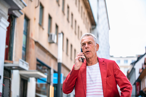 Low angle front view close-up of Caucasian man in early 60s wearing smart casual clothing and pausing to take phone call while walking in Ljubljana.